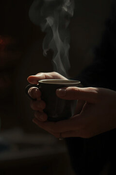 Cinematic detail of hands holding a steaming ceramic cup, dramatic highlights tracing the fingers while soft mist curls upward, minimal background with low key atmosphere enhancing