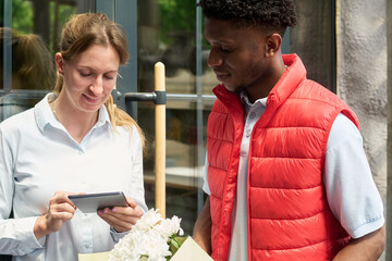 young woman showing digital tablet to Black young man standing together outdoors, both focusing on screen, bouquet of flowers visible in foreground