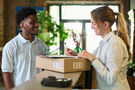 Young Black man delivering package to middle aged woman signing document at counter, both standing indoors with plants in background, engaging in delivery transaction