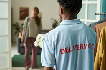 Young Black man wearing delivery company uniform entering modern home holding bouquet of flowers, young woman standing in background greeting delivery worker
