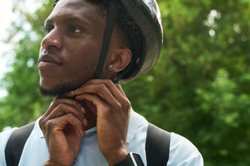 Young Black man fastening bicycle helmet strap outdoors, wearing backpack and smartwatch, looking away from camera with thoughtful expression, surrounded by green foliage