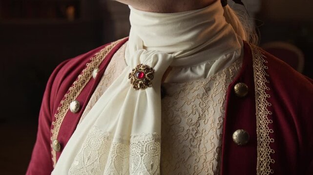 Medium close-up of anonymous cleanly shaven, fashionably dressed 18th century gentleman in red frock coat, brocade vest, lace cravat with brooch, wig standing by window