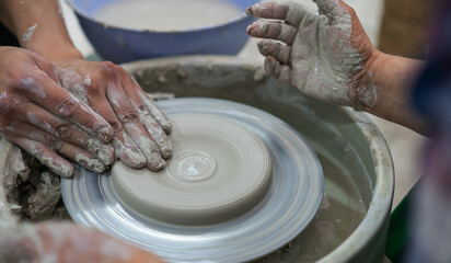 Close-up of hands covered in wet clay as two people collaboratively shape a pottery piece on a spinning wheel, emphasizing the teamwork and artistry involved in ceramic craftsmanship.