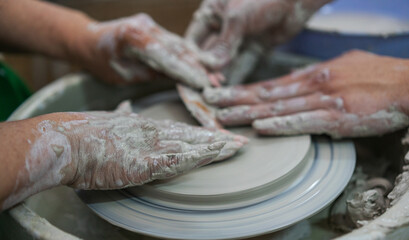 Close-up of hands covered in wet clay as two people collaboratively shape a pottery piece on a spinning wheel, emphasizing the teamwork and artistry involved in ceramic craftsmanship.