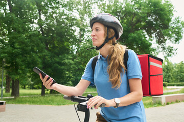 Young woman wearing helmet riding bicycle while holding smartphone and carrying insulated delivery backpack outdoors in park setting, focused on navigation or delivery task