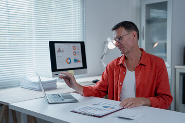Mature entrepreneur working with laptop and computer showing statistical graphs and charts, analyzing financial data and planning project strategy in modern office