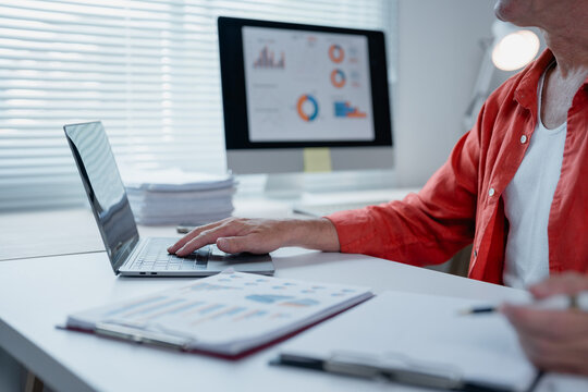 Senior businessman sitting at desk working with laptop and analyzing financial charts displayed on computer screen and printed documents, he is holding a pen and taking notes - Powered by Adobe