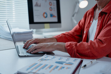 Businessman wearing a red shirt sitting at office desk and typing on laptop keyboard, analyzing financial statistics displayed on computer screen and working with charts and graphs