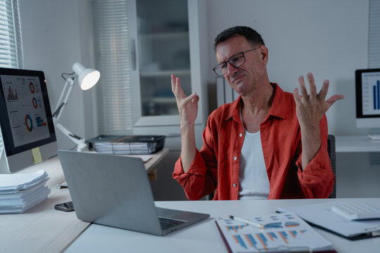 Stressed businessman sitting at a desk in an office at night, explaining complex ideas during a video call on a laptop, using hand gestures to emphasize points while managing pressure