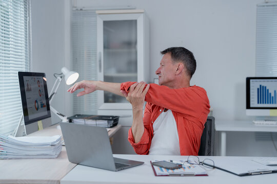 Mature office worker is stretching his arm while sitting at his desk, taking a break from working on laptop and desktop computer to relieve muscle tension and improve flexibility