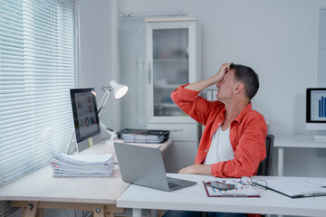 Businessman experiencing a headache while working on a laptop in his office, surrounded by paperwork and computer screens displaying graphs, indicating work stress and pressure