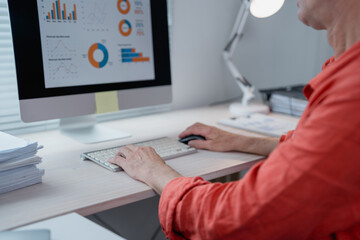 Businessman working at desk typing on a keyboard and using a mouse while analyzing financial charts and graphs displayed on a computer screen in a bright office