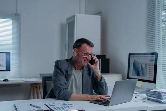 Senior businessman talking on phone and working on laptop in modern office, checking charts and graphs on computer screen, analyzing financial data and discussing business strategy