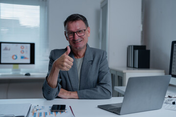 Fototapeta premium Senior business manager giving a thumbs up while sitting at a desk in a modern office, radiating positivity and satisfaction with work achievements