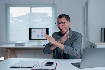 Businessman sitting at desk in office, showing stop gesture with crossed arms and open mouth, expressing disapproval or rejection, with laptop and documents on table
