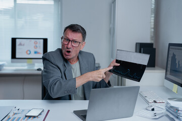 Furious mature businessman in eyeglasses pointing at declining sales chart showing financial crisis during video call with colleagues in office