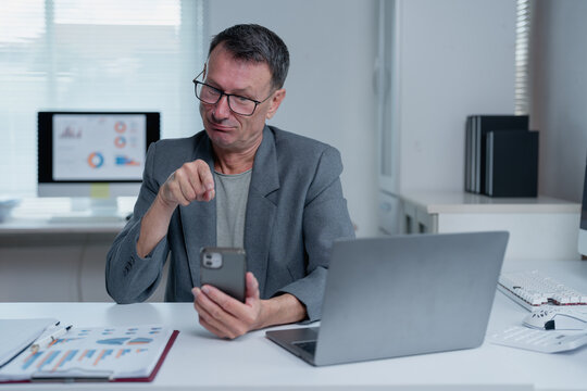 Businessman is sitting at his desk in the office, having a video call on his smartphone and pointing his finger at the screen, with a laptop and financial reports on the desk