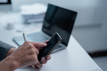 Businesswoman using smartphone and holding pen while working at office desk with laptop and documents in background, representing modern communication and multitasking in business environment