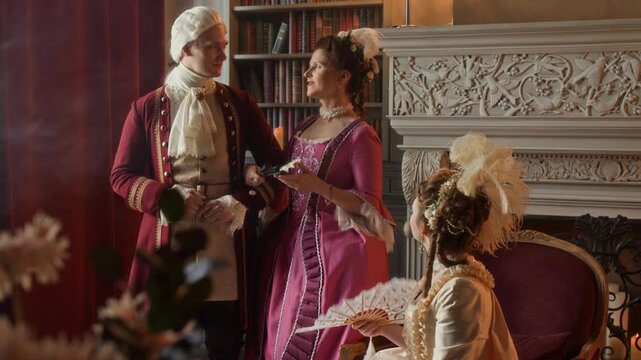 Medium full shot of aristocratic couple paying visit to duchess friend in 18th century manor, young Caucasian lady holding husband in surcoat by hand, chatting and gossiping while waving fans