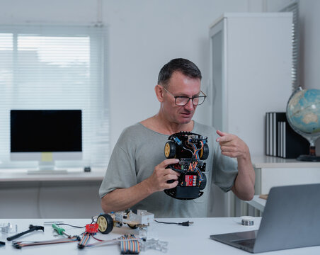 Robotics engineer explaining and showing robotic device during a video call on his laptop, sitting at his desk with electronic components and tools