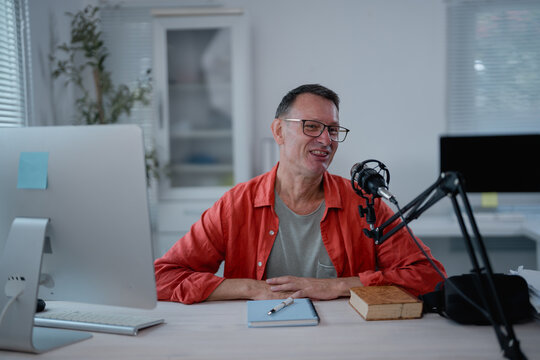 Middle-aged broadcaster smiling while hosting a radio show or podcast from a home studio, engaging with listeners through a professional microphone and computer setup