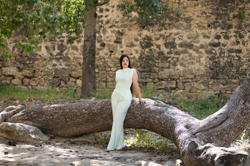 Young, beautiful, brunette woman in a white dress, posing looking at infinity leaning on the trunk of a fallen tree. Concept beauty, fashion, trendy, youth.