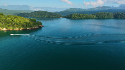 Aerial view of a boat slicing through the tranquil, turquoise waters surrounded by verdant islands and distant, hazy mountains, Paraty, State of Rio de Janeiro, Brazil.
