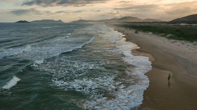 Aerial view of the shoreline where foamy waves crash onto the sandy beach with a lone figure enjoying the sunset, Paraty, State of Rio de Janeiro, Brazil.