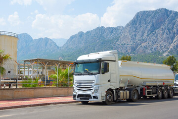 A long, white tanker truck is parked on the side of a road. Behind the vehicle is an industrial facility and a range of mountains in Antalya, Turkey, during the daytime