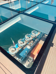 Unique perspective through a transparent glass panel revealing a waterfront area below. Circular seating with blue umbrellas lines a dock, accompanied by palm trees, kiosks, and an Italian flag