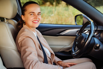 A cheerful woman in a tan blazer smiles directly at the camera while sitting in the driver's seat of her car.