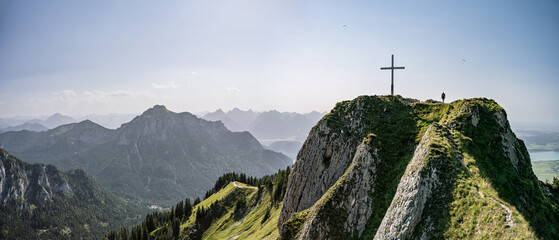 Hiker standing on the summit of Branderschrofen on a hot summer day, with Säuling and Füssen with its lakes in the background.