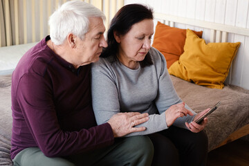 Old couple using a phone together while relaxing in the kitchen at home. Elderly man and woman spending leisure time browsing online with a smartphone. Happy senior married couple typing, learning