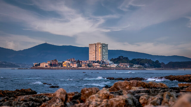 The Island of Toralla and Its Tower, Seen from the Coast. At the center of the island stands a tall, modern apartment tower, surrounded by smaller, colorful houses.