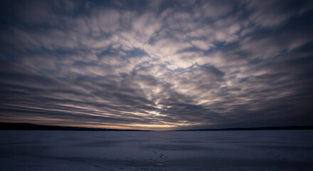 Fototapeta premium Expansive cloudy sky over a flat snowy landscape at dusk with a horizon glow and subdued colors