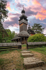 Fototapeta premium A traditional wooden church with a tall shingled tower stands surrounded by trees and a rustic fence under a colorful sunset sky, creating a serene and picturesque rural scene.