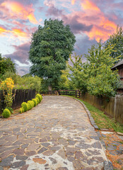 A rustic stone path in a countryside village with wooden fences, lush greenery, and a colorful sunset sky.