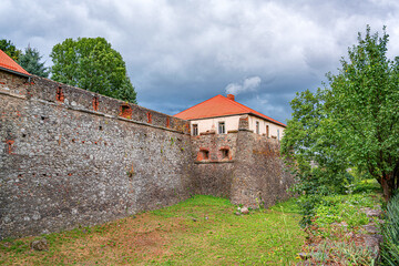 A historic medieval castle and its stone wall stand under a dramatic, cloudy sky, surrounded by green grass and trees. © Denis Rozhnovsky