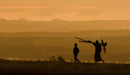 Bogura, Bangladesh - 10 November 2016: View of two silhouetted figures, one bearing tools, traversing an open field beneath a hazy, golden sky.