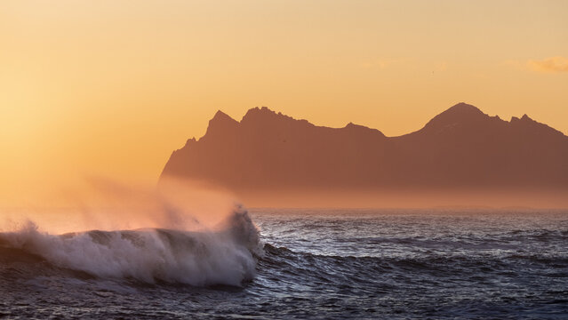 View of the dark waves crash against the shore beneath a fiery golden sky, silhouetting the jagged peaks of Vestmannaeyjar, Iceland.