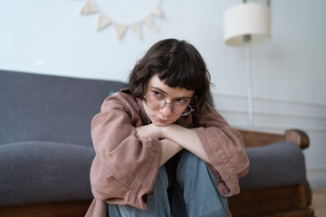 Unhappy teen girl sitting on floor devastated thinking about trouble, broken heart. Upset frustrated teenager being bullied or harassed online. Young woman have bad mood due to hormones in period. 