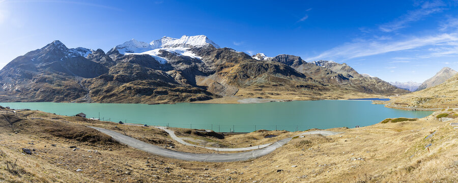 View of vibrant turquoise waters mirroring the snow-capped mountains under a clear azure sky, with winding path cutting through golden fields, Samedan, Grisons, Switzerland.
