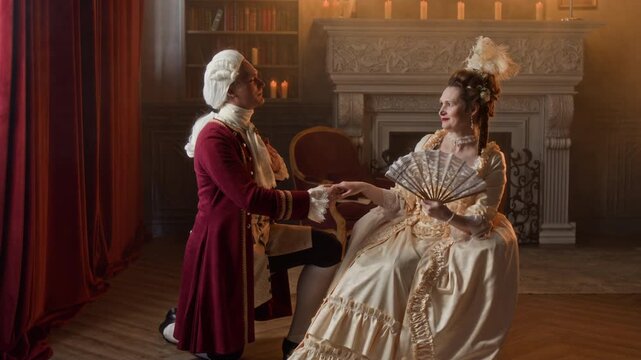 Full shot of elegant young Caucasian lord in red frock coat, breeches and wig standing on knee in front of fashionably dressed noble lady, confessing love and asking for her hand in marriage