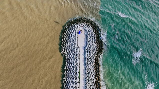 A long stone breakwater juts from the sandy shore into vast blue Loc An, creating calm shallow areas and vivid water patterns beneath a bright clear sky.