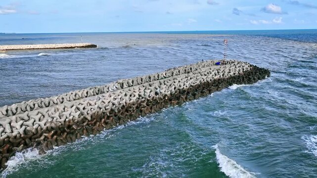 A long stone breakwater juts from the sandy shore into vast blue Loc An, creating calm shallow areas and vivid water patterns beneath a bright clear sky.