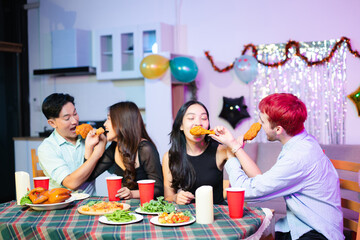 Playful moment as joyful friends laugh and feed each other fried chicken at a festive indoor dinner table, celebrating together with food, fun, and red party cups.