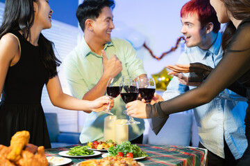 Closeup of smiling friends toasting with red wine at a lively indoor party table, sharing food, laughter, and joyful moments in a cozy