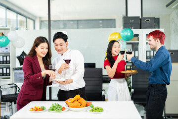 Cheerful coworkers share laughs as one playfully feeds a skewer during a festive office lunch with wine, food, and laughter, celebrating together in a modern workplace.