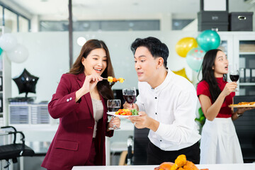 Cheerful coworkers share laughs as one playfully feeds a skewer during a festive office lunch with wine, food, and laughter, celebrating together in a modern workplace.