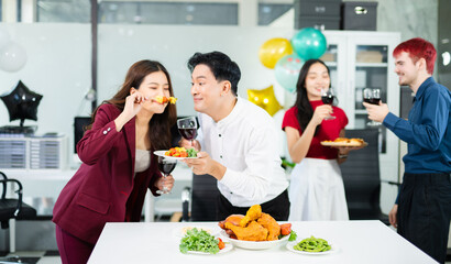 Cheerful coworkers share laughs as one playfully feeds a skewer during a festive office lunch with wine, food, and laughter, celebrating together in a modern workplace.